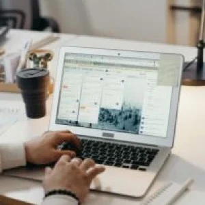 Hands typing on a laptop in a modern office setting with notes and task management software visible on the screen.